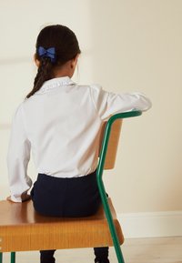 Cămașă albă cu guler fronsat, purtată cu pantaloni negri închis, așezată pe un scaun din lemn cu un cadru metalic verde.
