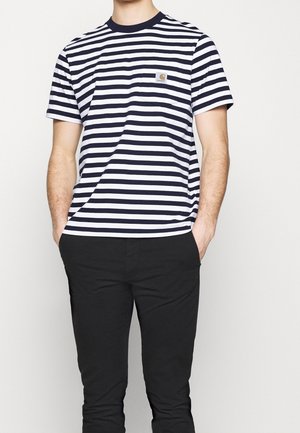 Mand iført en sort og hvid kortærmet t-shirt med horisontale striber, brystlomme og sorte bukser, hænderne i lommerne.