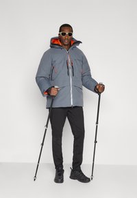 Šedá péřová bunda s oranžovým vnitřkem, zapínáním na zip vpředu a zipovými kapsami. Černé kalhoty a trekové hole doplňují outfit.