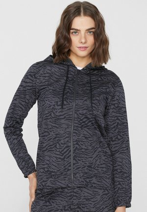 Junge Frau trägt einen schwarz-grau gestreiften Tiger-Zip-Hoodie mit Kapuze und Kordeln, steht mit einer Hand in der Hüfte.