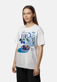 Žena v bielom tričku s modrou grafikou zobrazenou na miske farebného ovocia a s textom "Fichi Viola" a "Tempo de relax."