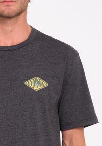 T-shirt gris à manches courtes en tissu doux, avec un logo jaune et vert en forme de diamant sur le côté gauche de la poitrine.