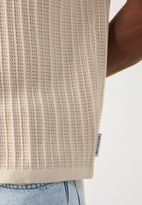Beige knitted sweater with a textured design, vertical ribbing, and a label on the side. The material appears soft and breathable.