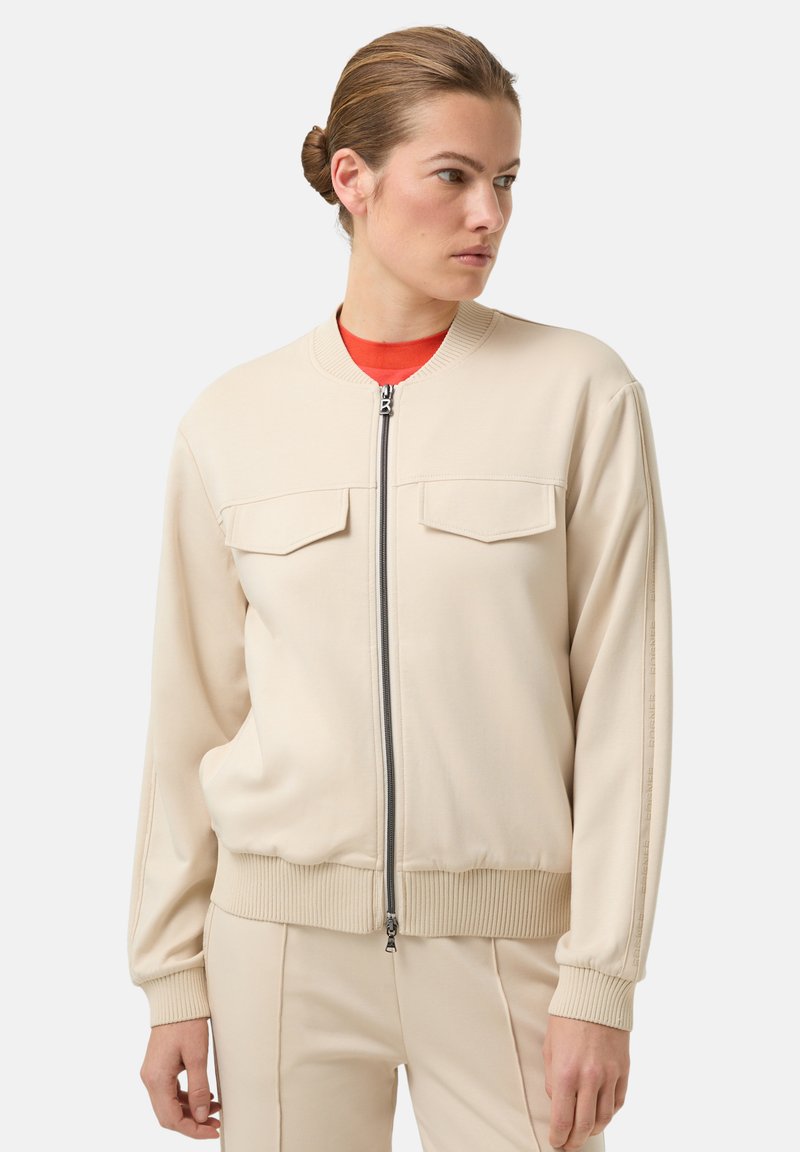 Béžová bomber bunda z měkké látky, s předním zipem, dvěma náprsními kapsami, žebrovanými rukávy a kulatým výstřihem.