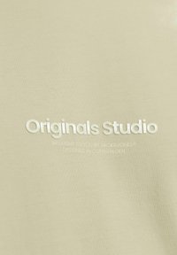 Helles gelbes Baumwoll-T-Shirt mit dem Text "Originals Studio" in Weiß. Enthält zusätzlichen Text über die Marke und den Designstandort.