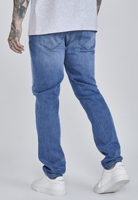 Blue denim jeans with a relaxed fit, visible stitching, and two back pockets. Worn with white sneakers.