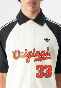 White and black short-sleeve polo shirt featuring a buttoned collar, red "Originals" graphic, and the number "33" printed in orange.