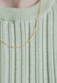 Colier din aur așezat pe un pulover ribatat de culoare verde deschis. Lanțul are un design delicat, răsucit și o textură fină.