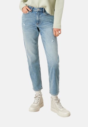 Henkilö pukeutunut vaaleansinisiin kuluneisiin farkkuihin, beigeihin platvormitennareihin ja vaaleanbeigeen pitkähihainen yläosa käsivarsi taskussa.