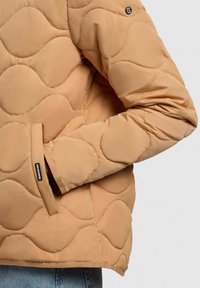 Giacca imbottita beige con motivo a onde, dotata di tasca laterale e etichetta con logo nera. Tessuto liscio e vestibilità su misura in evidenza.