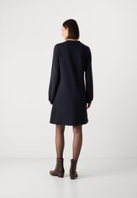 Marineblauwe jurk met lange mouwen en een ontspannen pasvorm, met een gebogen zoom en subtiele stiksels. Draagt men met bruine enkellaarsjes en doorschijnende panty’s.