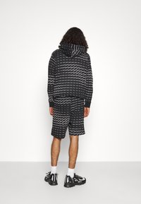 Person med lockigt hår iklädd svart hoodie och shorts med vita streckade linjer och mönster med varumärket, svarta sneakers, som står vänd mot en vit vägg.