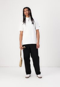 Tricou alb din bumbac cu un grafism negru pe piept, asortat cu pantaloni negri largi și adidași albi cu tălpi maro. Ținând o geantă bej.