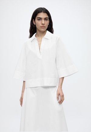 Mujer con cabello largo y oscuro que lleva una blusa blanca suelta con mangas anchas y una falda blanca, de pie frente a un fondo claro y liso.