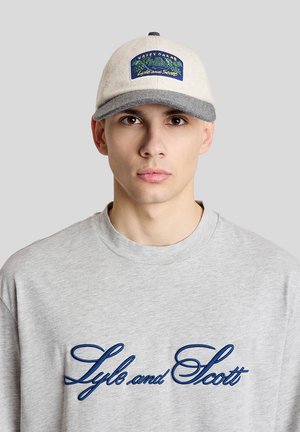 Jonge man met een grijze trui met ronde hals met de tekst "Lyle and Scott" en een beige en grijze pet met een kleurrijke patch aan de voorkant.
