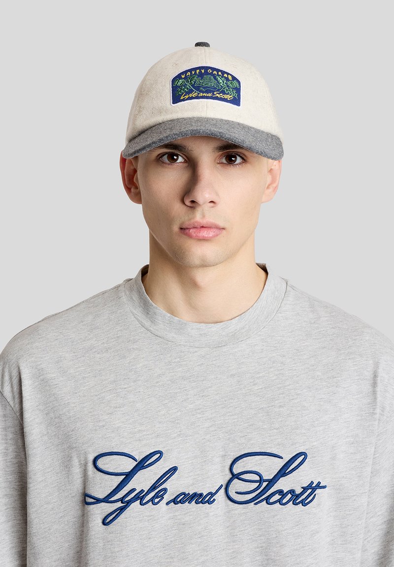 Νέος άνδρας που φορά γκρι φούτερ με στρογγυλή λαιμόκοψη και το κείμενο "Lyle and Scott" και μπεζ και γκρι καπέλο με πολύχρωμο σήμα στο μπροστινό μέρος.