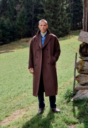 Homme portant un long manteau marron debout sur l’herbe près d’une structure en bois avec des arbres en arrière-plan, les mains dans les poches, regardant devant.