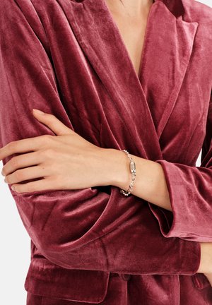 Blazer de terciopelo color burdeos con solapas anchas y textura suave. Pulsera plateada con pequeñas piedras y un cierre alargado en la muñeca.