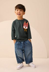 Ciemnozielona koszula z długim rękawem z motywem kieszeni w kształcie lisa, połączona z luźnymi niebieskimi dżinsami i białymi trampkami.