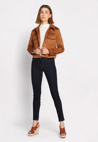 Bruine suedine cropped jas met knopenclosure, vier zakken, gedragen over een lichte turtleneck en donkere slim fit jeans. Bruine wedge sandalen maken de look compleet.