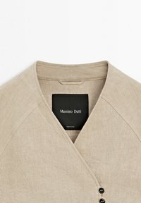 Beige linnedragt med V-udskæring og sort label med teksten "Massimo Dutti". Skjorten har asymmetrisk forside og synlige syningsdetaljer.