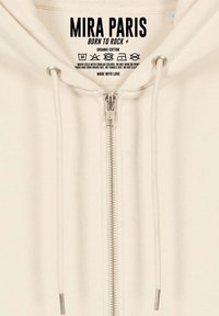 Felpa con zip di colore crema realizzata in cotone biologico. Presenta una zip argentata, coulisse e un'etichetta con logo sul collo.