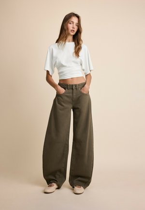 Jeune femme debout les mains dans les poches, portant un top court blanc, un pantalon large vert olive et des chaussures plates beiges.