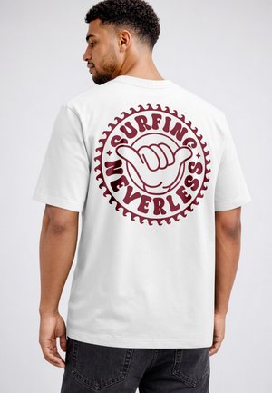 Man wearing white t-shirt with maroon "Surfing Neverless" text and shaka hand sign graphic on the back, facing sideways.