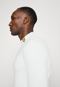 Long-sleeve white top made of stretchy material, featuring a black and yellow butterfly patch on the collar. Smooth, fitted design.