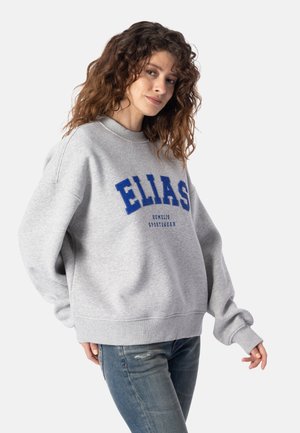 Ung kvinde med krøllet hår iført en grå sweatshirt med "ELIAS" i store blå bogstaver og falmede blå jeans, stående mod en ensfarvet baggrund.