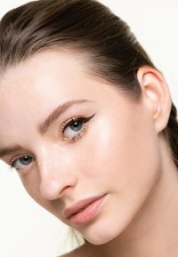 Nära bild av ett ansikte med slät hy, ljusa fräknar, blå ögon och tydlig eyeliner. Håret är slätt och bakåtkammat, vilket avslöjar ett öra.