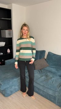 Γυναίκα με μακριά ξανθά μαλλιά στέκεται στο σαλόνι φορώντας ριγέ πουλόβερ, μαύρο παντελόνι και μπεζ ψηλοτάκουνα δίπλα σε καναπέ από μπλε κοτλέ ύφασμα.