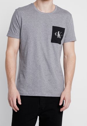 Graues Kurzarm-T-Shirt mit Rundhalsausschnitt und einer schwarzen aufgesetzten Tasche, die ein weißes Logo zeigt. Der Stoff wirkt glatt und lässig.