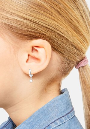 Orecchino a cerchio in argento con ciondolo a stella pendente, finitura in metallo liscia e texture lucida, visto di lato accanto a una coda di cavallo.