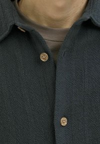 Donkergroen button-up shirt van een gestructureerde, ribgebreide stof. Bevat natuurlijke houten knopen en een kraagdesign over een beige onderhemd.