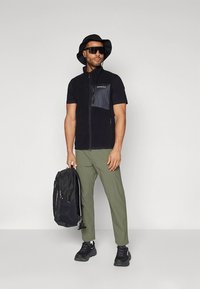 Zwart fleecejack met korte mouwen en een blauwe borstzak, groene broek, zwarte sneakers, een rugzak en een zwarte bucket hat. Donkere zonnebril op.