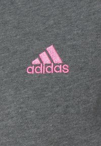 Tissu en coton gris présentant un logo Adidas rose brodé avec trois bandes diagonales en haut, positionné sur le côté gauche.