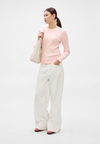 Ružový sveter s vrkočovým vzorom, široké biele nohavice, béžová kabelka a dvojfarebné topánky s ružovými detailmi. Modelka stojí pred jednoliatym pozadím.