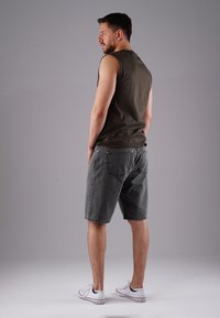 Man standing sideways wearing a dark sleeveless shirt, gray denim shorts, and white sneakers against a plain gray background.