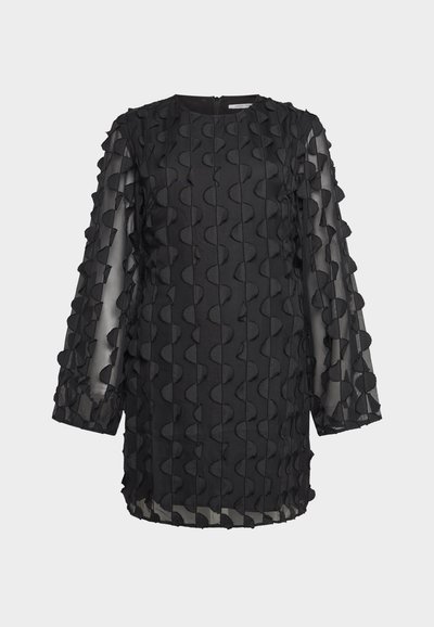 Tunique noire à manches longues et transparentes, ornée de détails en tissu ondulés en relief, sur un fond uni.