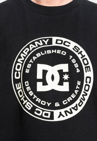 Černé bavlněné tričko s bílým kruhovým grafickým vzorem s nápisem "DC Shoe Company" a "Založeno 1994", umístěné na hrudi.