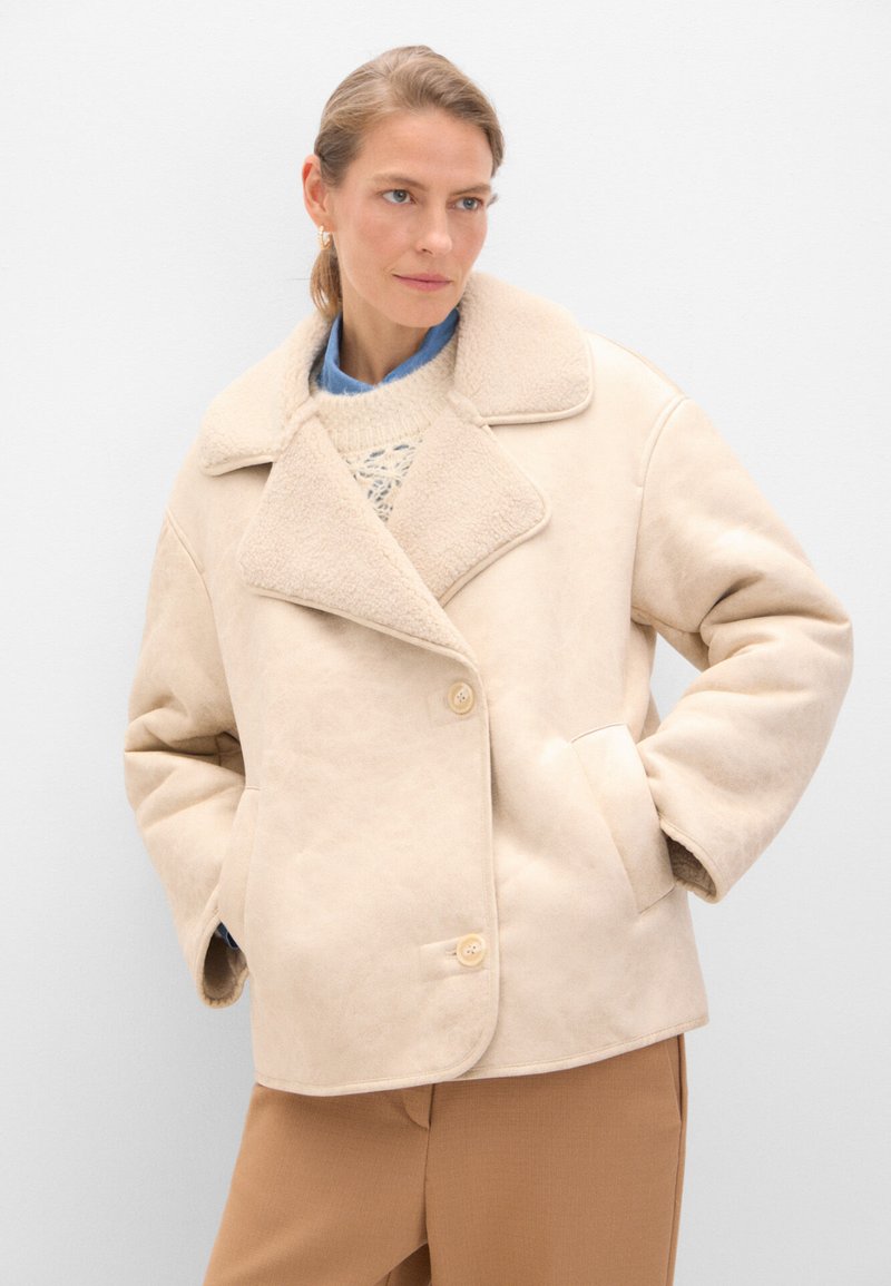 Giacca in shearling beige con ampio colletto, design doppiopetto, due tasche frontali e chiusura a bottoni, indossata sopra un leggero maglione.