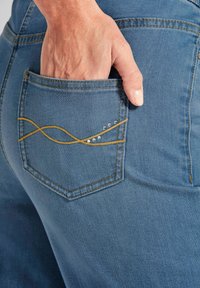 Jeans aus Denim in hellblauer Farbe. Verfügt über eine Gesäßtasche mit gelbem Stichmuster und dekorativen metallischen Perlen.