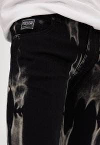 Svarta denimjeans med en urtvättad, sliten mönstring. Versace Jeans Couture-märke på midjebandet. Femfickorsdesign med metallhardware.