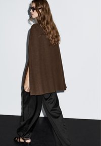 Poncho a coste marrone con spacchi laterali abbinato a pantaloni neri in satin e sandali scuri, con capelli lunghi e mossi e occhiali da sole.