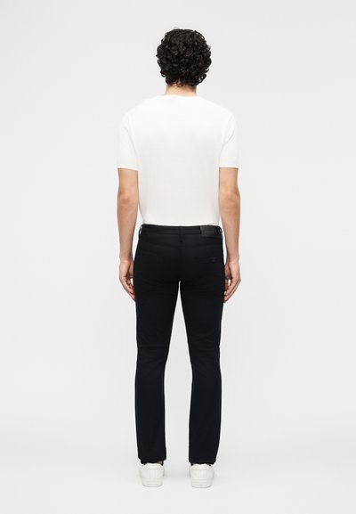 Άνδρας με σγουρά σκούρα μαλλιά, φορώντας απλό λευκό κοντομάνικο πουκάμισο, στενά μαύρα παντελόνια και λευκά αθλητικά παπούτσια, στέκεται με την πλάτη προς τα εμπρός.