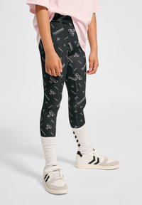 Svarta leggings med ett upprepande mönster av vitt logotyp, med en slät textur. Kombinerat med vita skor och randiga strumpor.