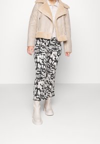 Béžová bunda s podšívkou z beránka a límcem, spárovaná s vzorovanými černobílými širokými kalhotami a světle šedými kotníkovými botami.