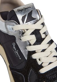 Black sneaker featuring a suede and nylon upper, silver accents, white laces, and logo details on the tongue and side.