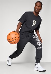 Juoda Nike marškinėliai su dideliu šviesiai pilku "9" logotipu, derinami su juodomis sportinėmis kelnėmis, ant kurių pavaizduoti balti krepšinio piešiniai ir balta Nike ženklinimas.